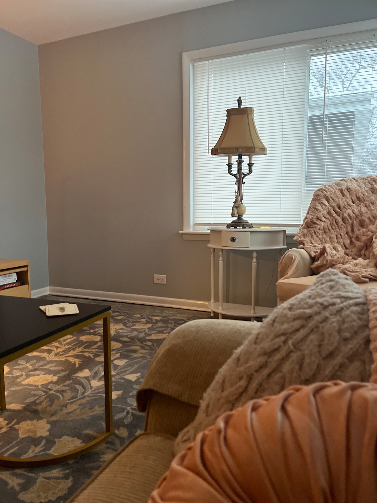 A cozy living room area features a plush sofa draped with a soft blanket, accompanied by a small side table and a stylish lamp. Natural light filters through partially closed blinds, creating a calming environment. A textured area rug adds warmth to the space.