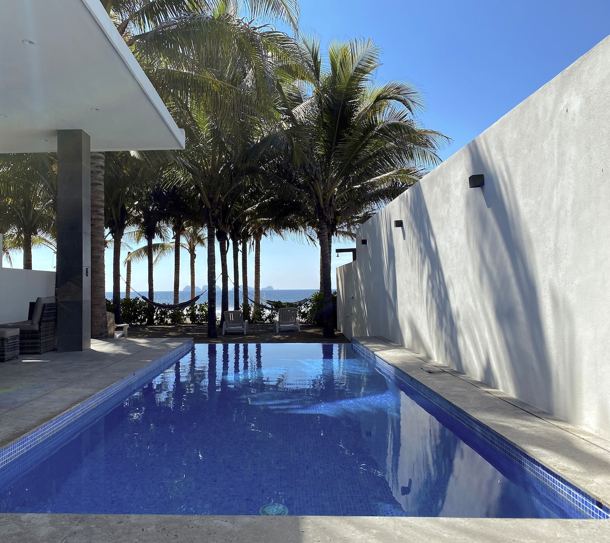 An infinity pool reflects the clear blue sky, surrounded by palm trees providing shade. The smooth stone deck contrasts with the vibrant water, creating a serene space for relaxation. White walls frame the area, enhancing the openness and connection to the natural surroundings.