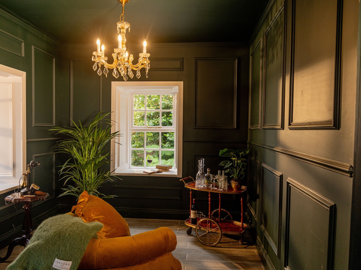 A lounge area features dark green paneled walls, complemented by a classic chandelier with warm lighting. A plush orange armchair is positioned beside a window, with a view of greenery outside. A vintage cart displays glassware, adding a touch of elegance to the space.