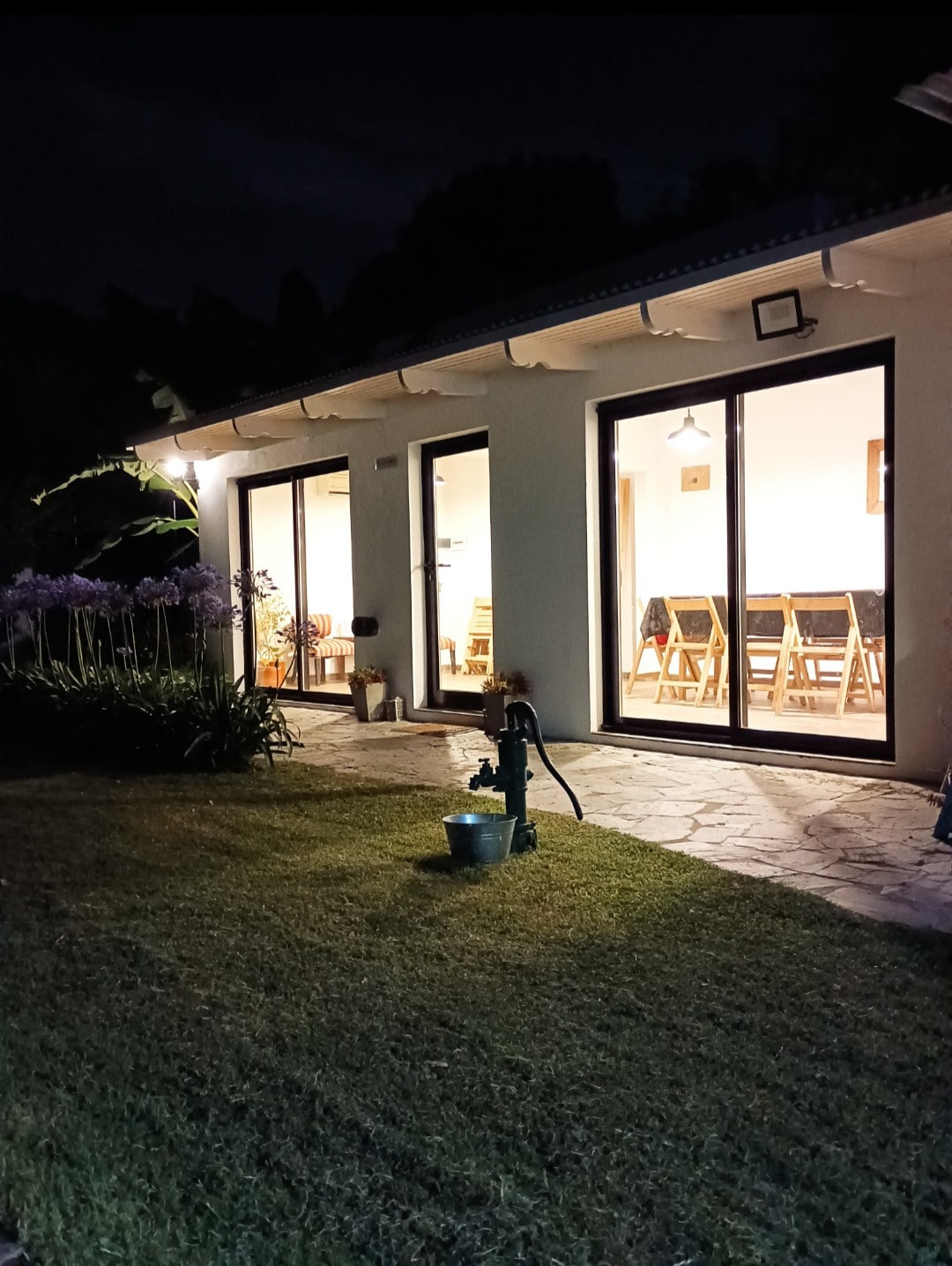 The exterior showcases large glass doors leading into a well-lit interior, framed by lush greenery and a manicured lawn. A garden area features flowering plants, and the scene is set in the evening, with soft lighting enhancing the inviting atmosphere.
