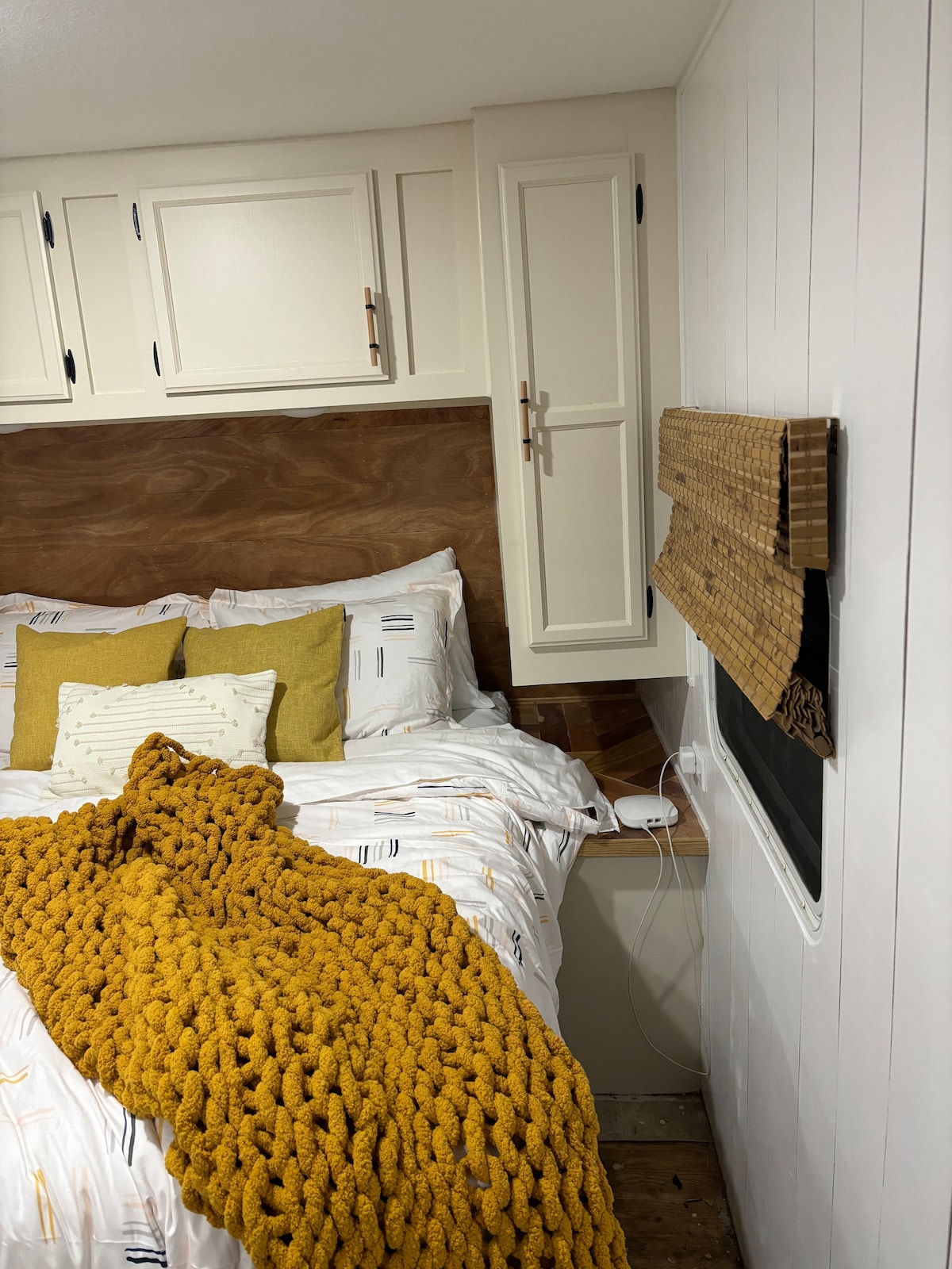 A cozy sleeping area features a spacious bed with white bedding accented by mustard yellow throw pillows and a chunky knit blanket. Natural light filters through a woven shade on a nearby window, enhancing the wood-paneled headboard and minimalist decor.