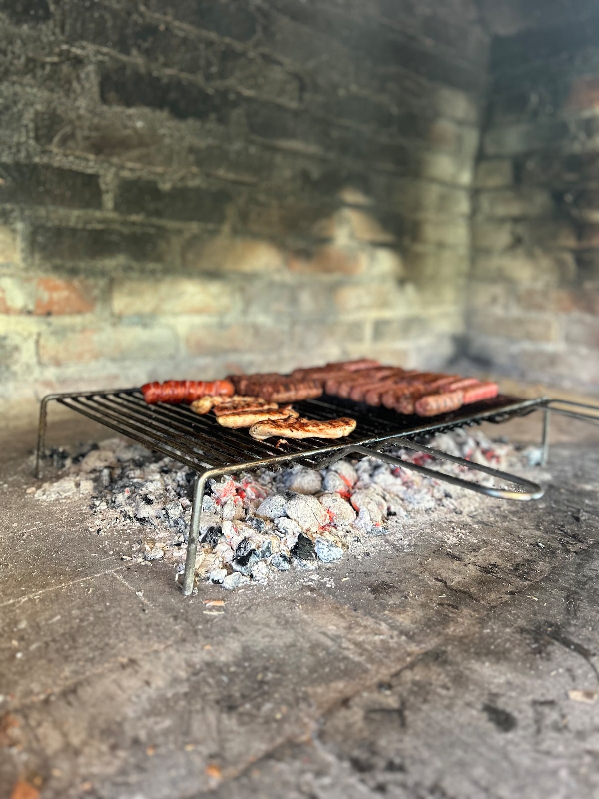 A grill set over glowing embers displays various meats sizzling, including sausages and steaks. The surrounding brick wall provides a rustic backdrop, creating an inviting cooking space for outdoor gatherings.