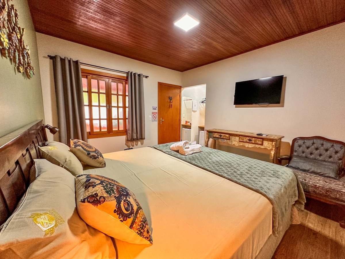 A spacious bedroom features a large bed adorned with decorative pillows and a cozy throw. A seating area is positioned next to a window dressed with gray curtains. The room includes a modern flat-screen TV mounted on the wall and an en-suite bathroom visible through an open door.