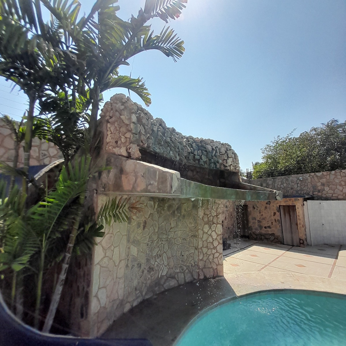 A natural stone structure features a cascading water feature, complementing the pool area. Lush green foliage surrounds the site, adding to the serene atmosphere. Bright sunlight illuminates the scene, highlighting the textures of the stone and the clarity of the water.