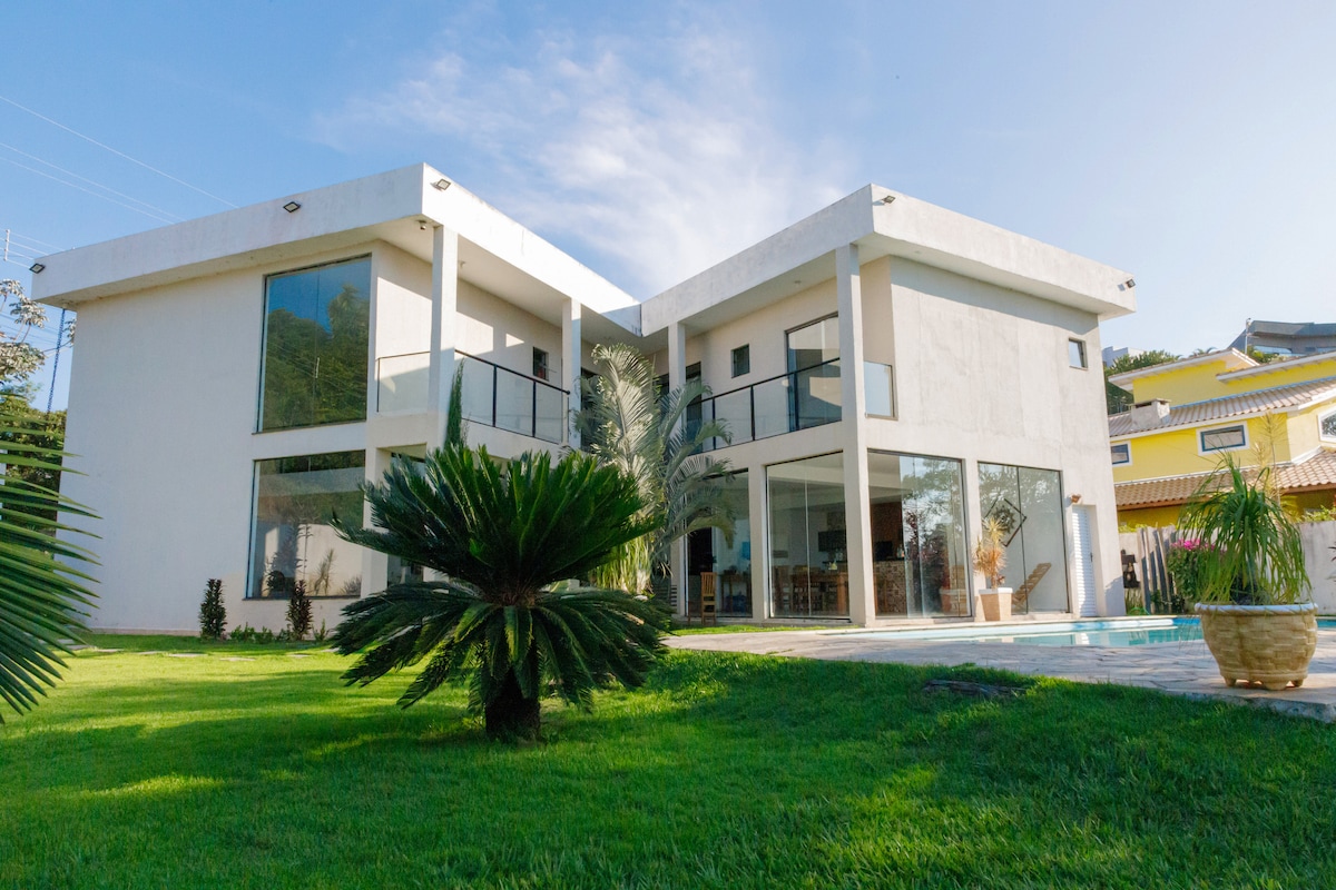 A modern two-story house is set amidst lush green grass, featuring large windows that allow natural light to fill the interiors. Balconies are present on each floor, providing outdoor views, while a palm tree enhances the inviting landscape.