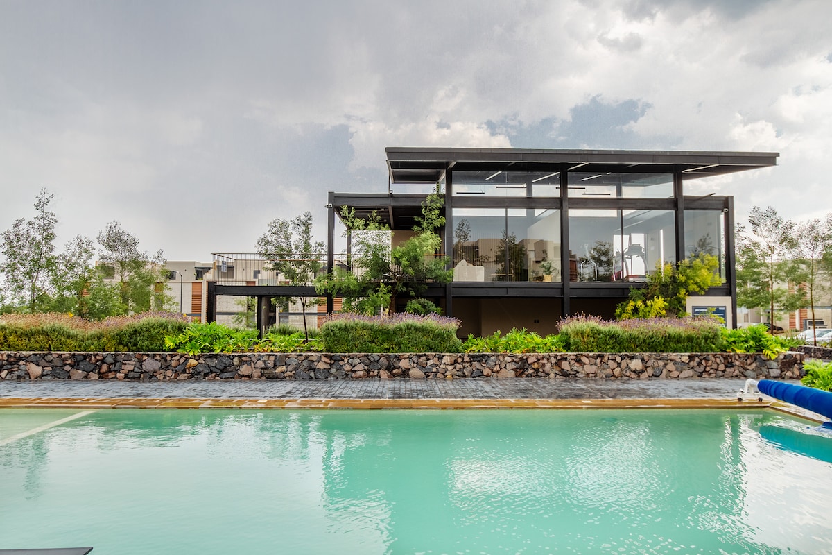 A modern two-story house is showcased beside a tranquil swimming pool. The structure features large glass walls, allowing for natural light and views of the landscaped surroundings. Lush greenery and decorative plant beds enhance the outdoor ambiance.