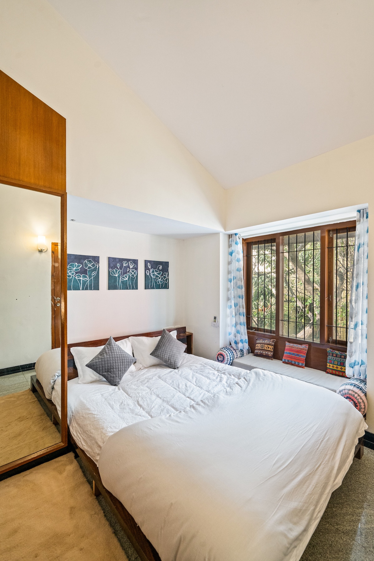 A spacious bedroom features a large bed with soft bedding and decorative throw pillows. Natural light enters through large windows framed with colorful curtains, creating a serene ambiance. An elegant mirror reflects the comfortable space, enhancing the overall brightness.