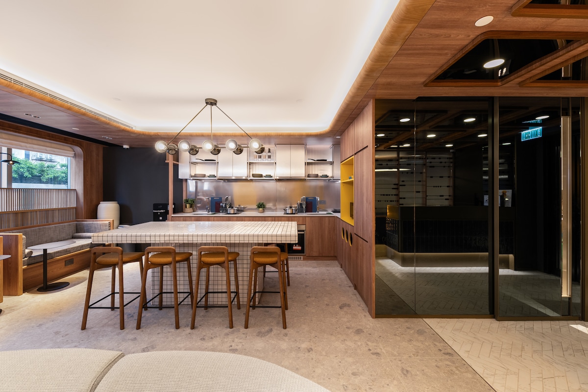 A modern kitchen area exhibits sleek cabinetry and a spacious island, complemented by a stylish light fixture above. Wooden bar stools are arranged around the island, and large windows allow natural light to fill the space, blending indoors with the view of greenery outside.