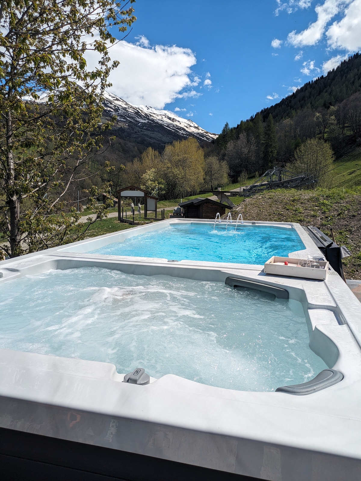 An outdoor spa area features a bubbling hot tub alongside a larger swimming pool, surrounded by a scenic landscape of green hills and snow-capped mountains. Clear blue skies are visible above, enhancing the tranquil setting.