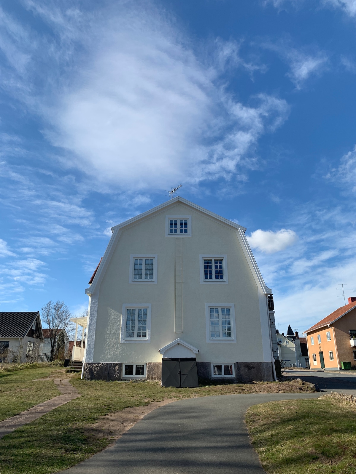 The exterior of a two-story house is depicted, featuring a simple facade with large windows on each level. The entrance is marked by a dark door, framed by light-colored walls. A clear blue sky with scattered clouds serves as the backdrop, enhancing the location's charm.