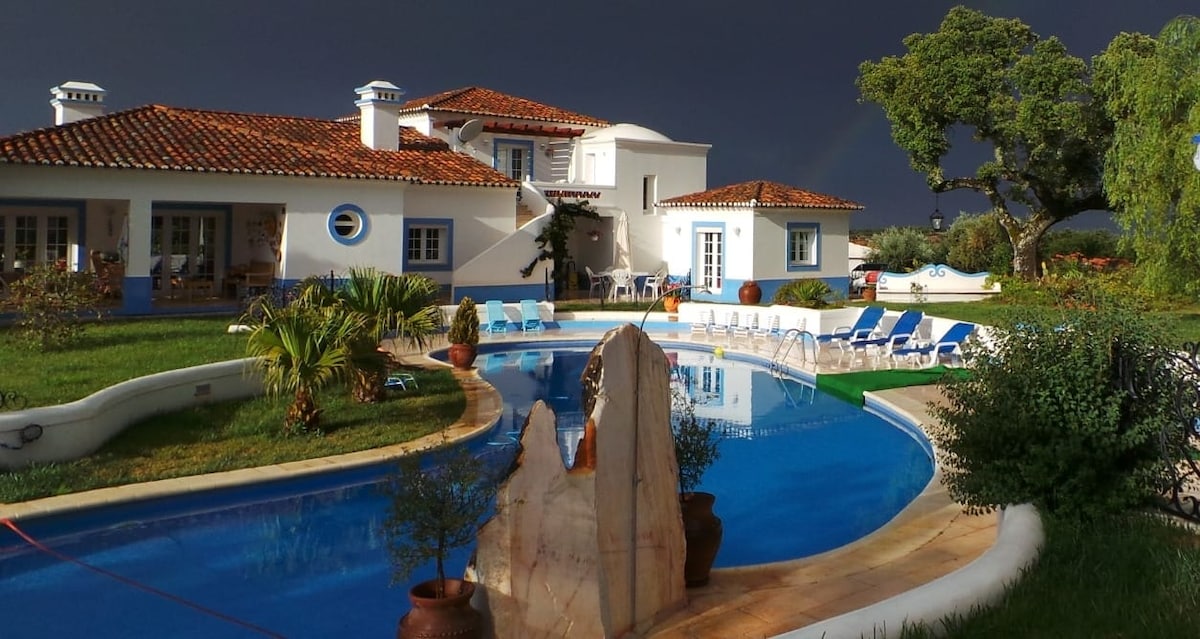 A serene outdoor area features a winding pool surrounded by neatly arranged lounge chairs. Lush greenery, including palm trees and potted plants, enhances the setting. The white exterior of the building is complemented by a terracotta roof, creating a harmonious blend with the natural surroundings.
