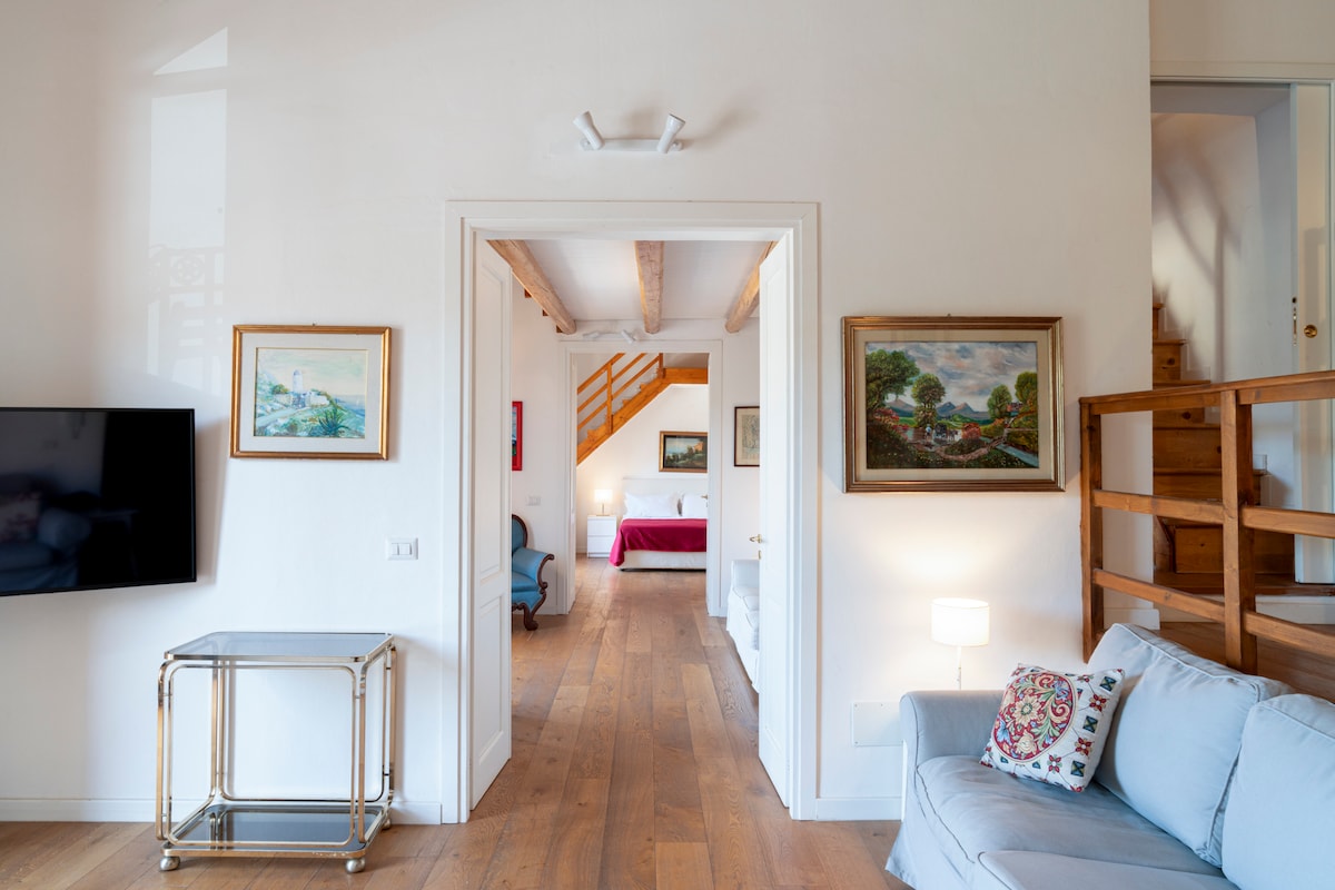 A bright hallway is shown, featuring light wooden floors and high ceilings. A comfortable sofa sits against one wall, while a TV is mounted on the opposite wall. Artworks adorn the walls, leading to an open space that includes a staircase and cozy bedroom area.