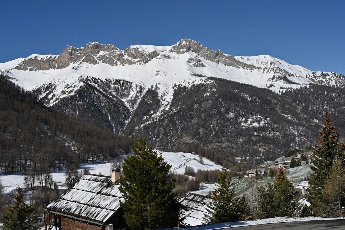 Snow-capped mountains rise majestically against a clear blue sky, showcasing a serene winter landscape. Pine trees frame the foreground, while a wooden structure with a rugged roof adds to the natural beauty. The distant valleys and slopes are blanketed in pristine snow.