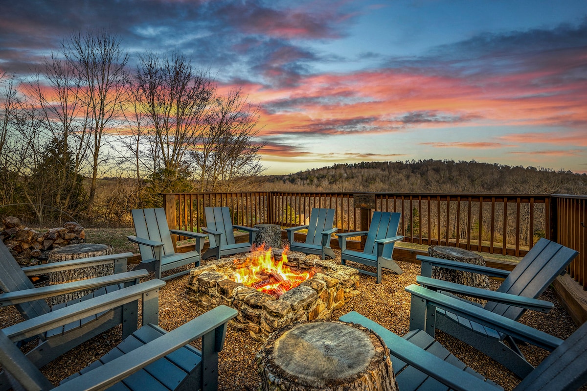 An outdoor fire pit area is surrounded by several blue Adirondack chairs, inviting relaxation. A stone circle encloses the fire, which flickers warmly against a backdrop of rolling hills and a colorful sky at sunset.