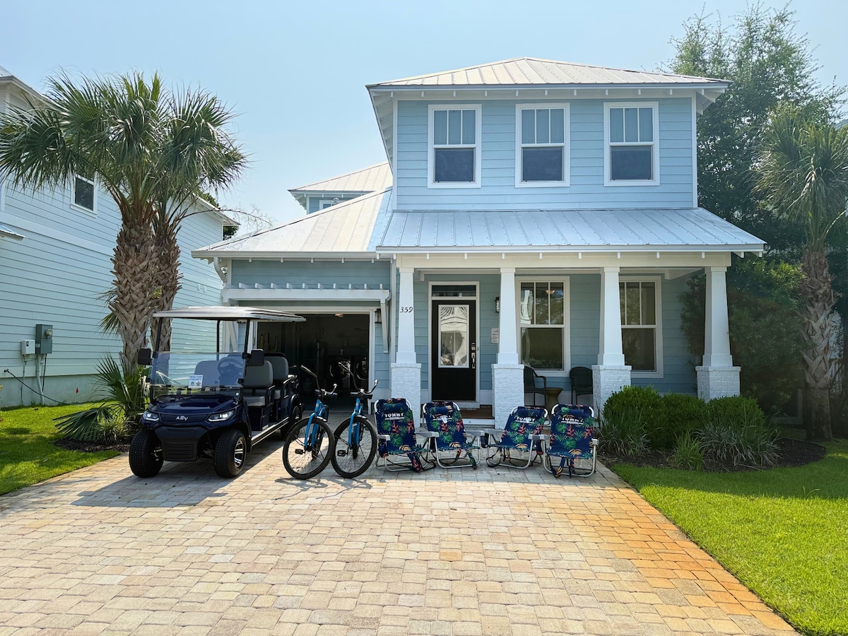 A charming coastal home features a light blue exterior with a welcoming front porch. A six-seater golf cart and two bicycles are positioned alongside six beach chairs on the paver driveway, bordered by palm trees and neatly maintained grass.