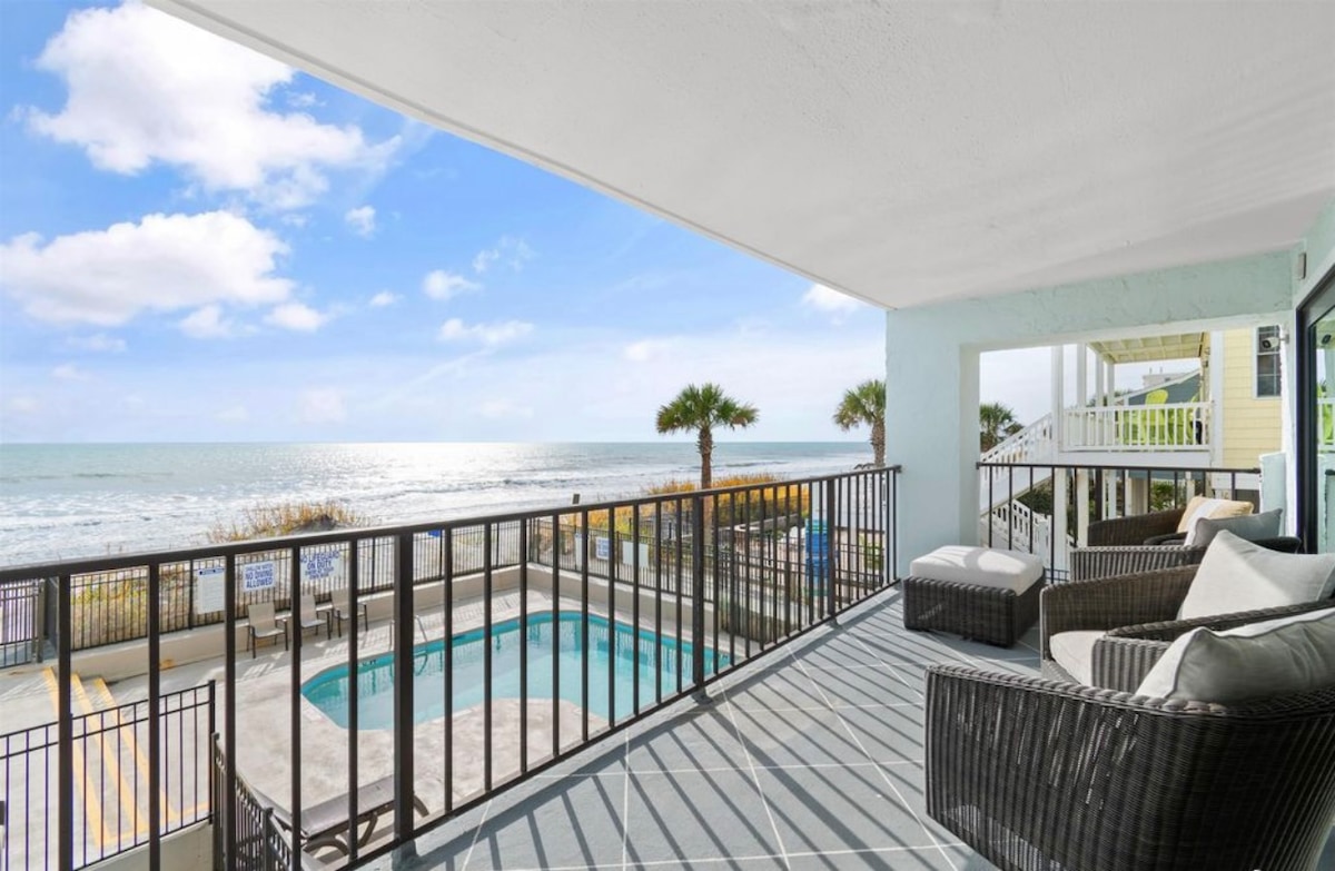 A spacious balcony offers direct ocean views, with a comfortable seating area featuring a wicker sofa and soft cushions. The shimmering pool below is framed by palm trees, creating a relaxing beachside atmosphere. Soft, natural light enhances the inviting space.