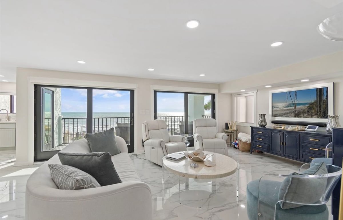 A bright living area features soft marble floors and large windows, providing views of the ocean. Comfortable seating includes a spacious sofa and recliners, complemented by a round coffee table and a media console with a large TV, enhancing the open ambiance.