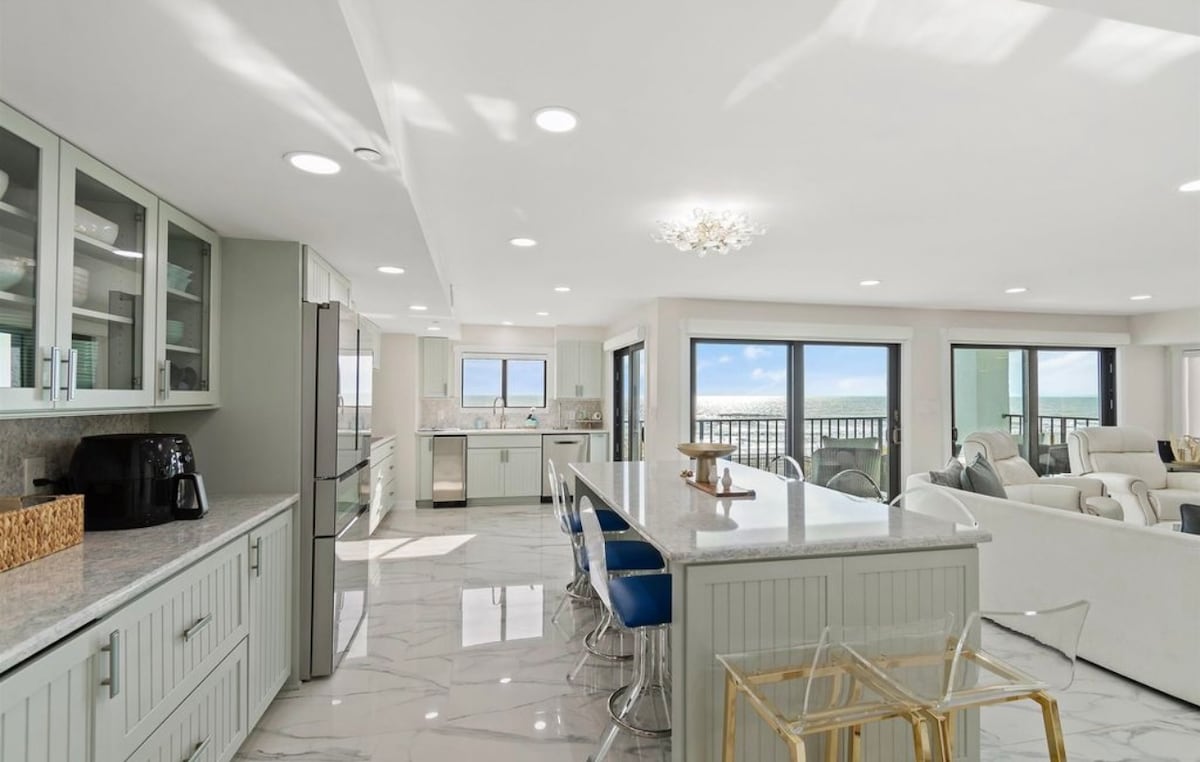 An open kitchen features modern appliances and a spacious island with seating. Light-colored cabinetry complements the marble floor, while large windows provide ocean views. The area is illuminated by natural light and a stylish chandelier, enhancing the inviting ambiance of the space.