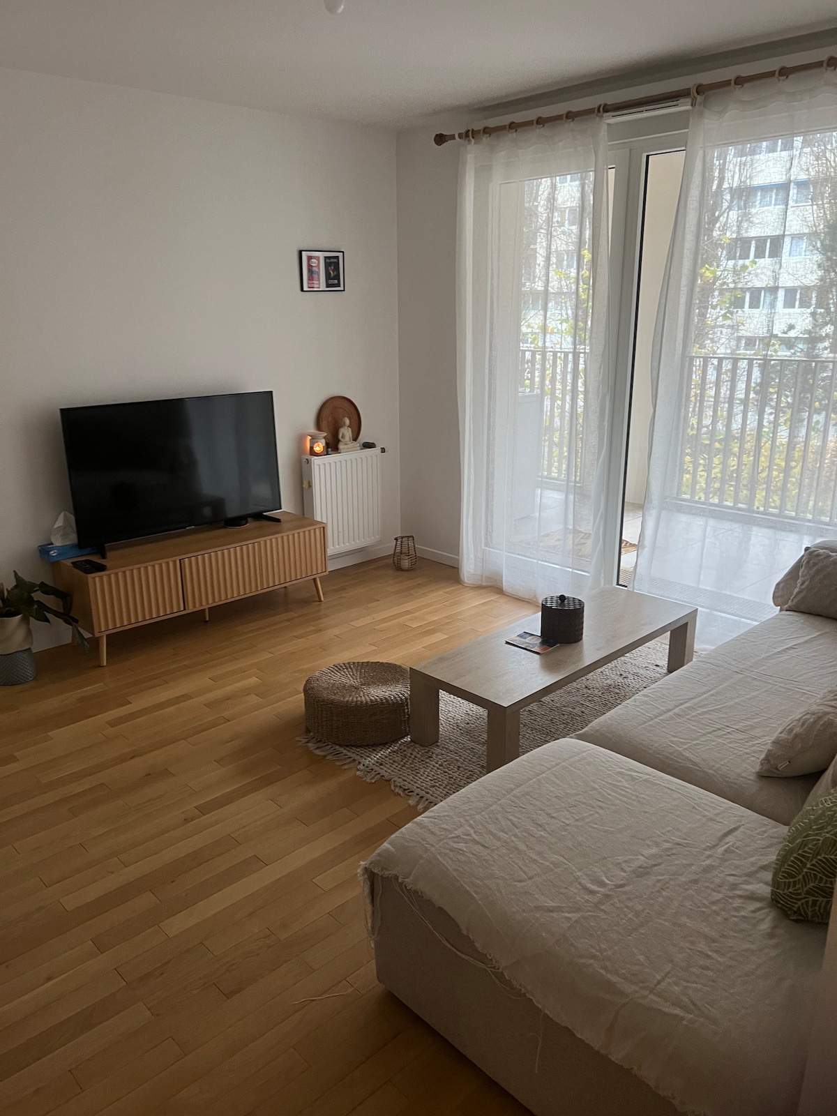 A cozy living area features a comfortable sofa and a low coffee table centered on an area rug. A television is placed on a light wooden media console, with gentle sunlight streaming through sheer curtains that frame a sliding glass door leading to a balcony.