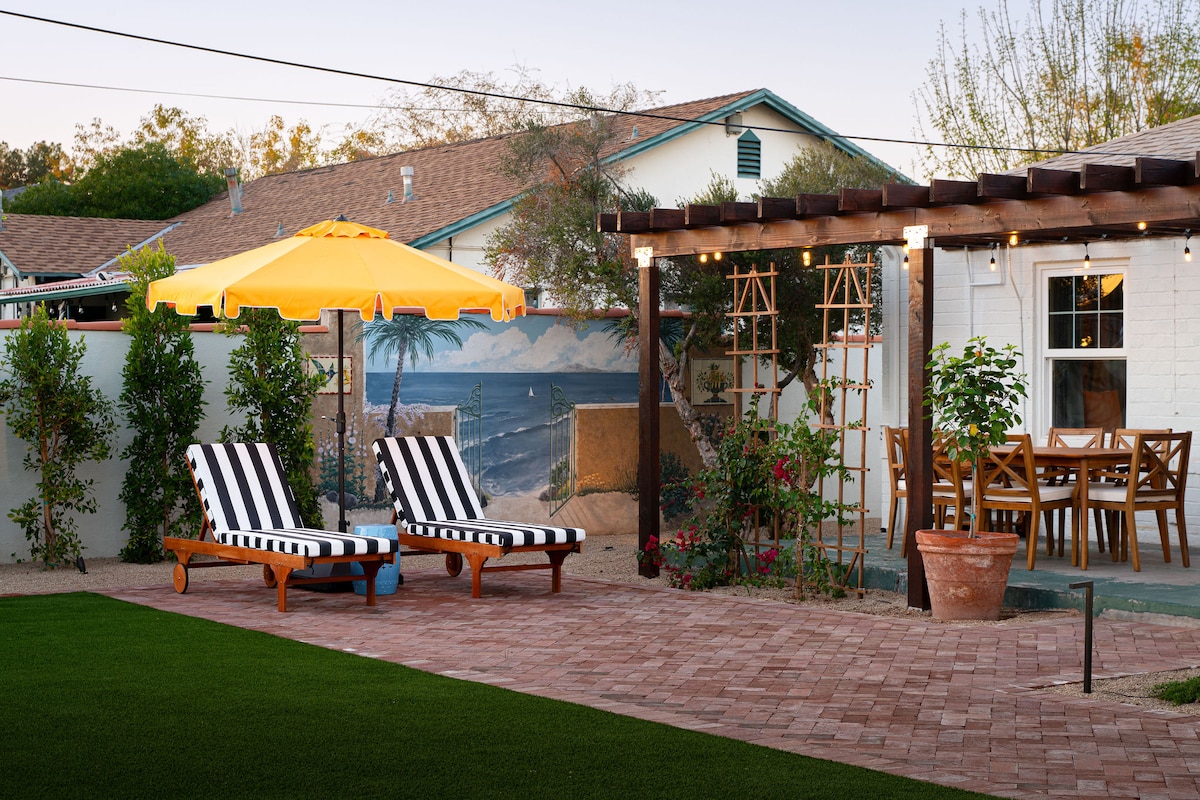 A vibrant backyard area features two striped sun loungers under a bright yellow umbrella, set on a manicured lawn. A rustic dining table with matching chairs is visible on a brick patio, surrounded by lush greenery and climbing plants, creating a calm outdoor retreat.