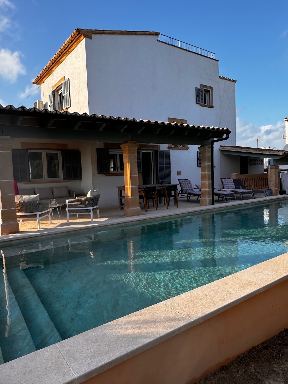 An inviting outdoor area features a swimming pool with clear water and steps leading into it. The pool is framed by a terrace with seating and a dining table under a covered patio, with additional outdoor loungers situated nearby.