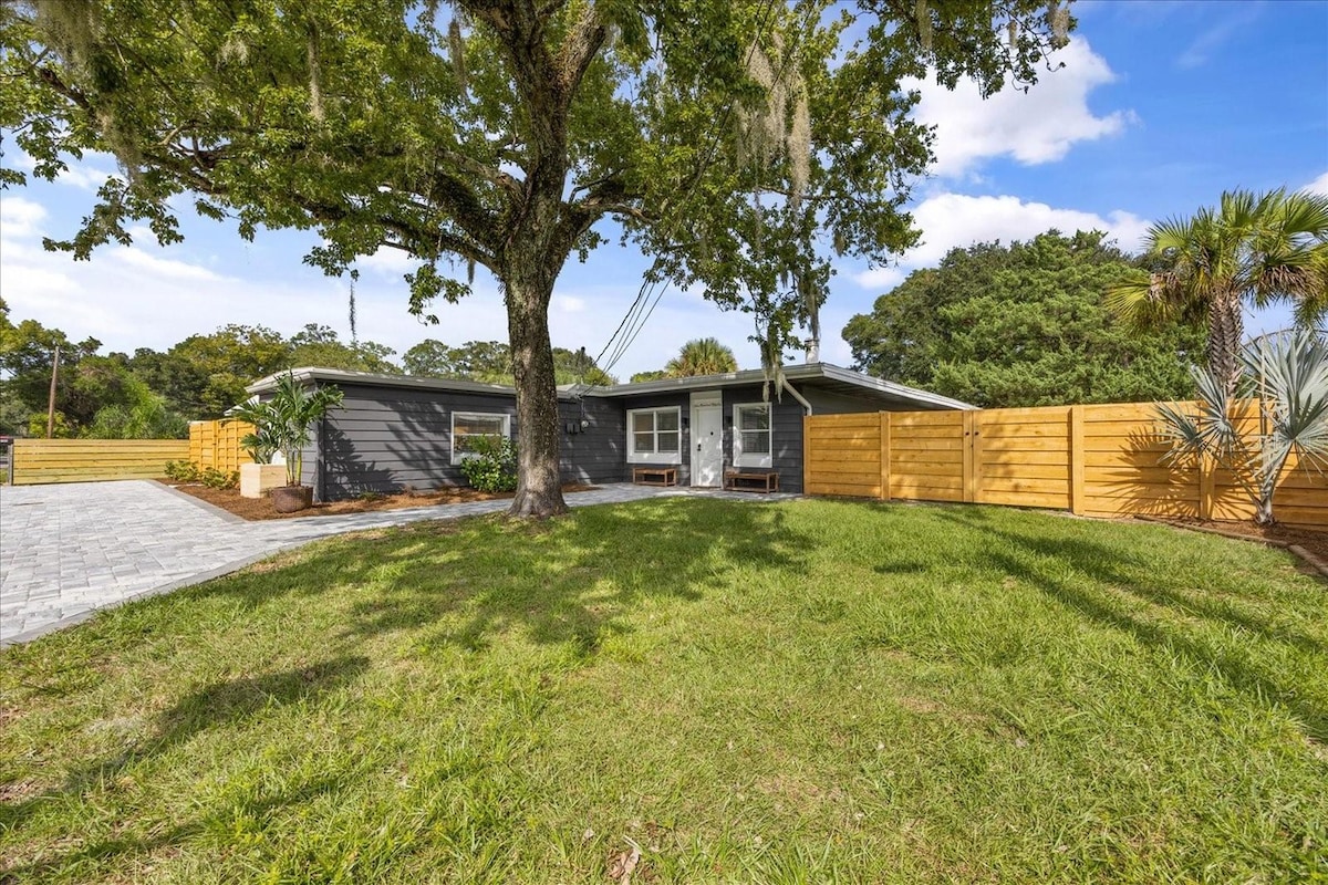 An expansive front yard features lush green grass and a large tree providing shade. The single-story house is partially visible, with a wooden fence surrounding the property. A stone pathway leads to the entrance, enhancing the inviting outdoor space.