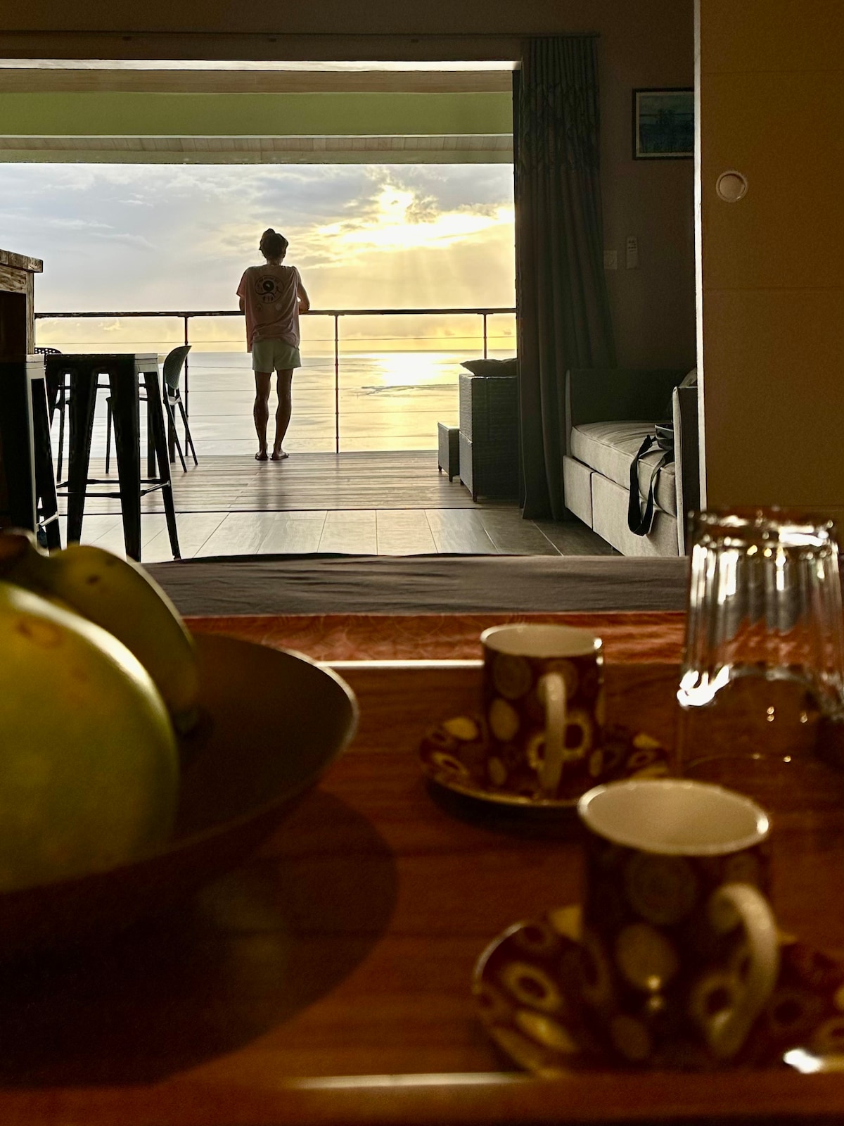 A serene interior view is captured, with a guest standing at a balcony overlooking the ocean and a sunset. A wooden table in the foreground holds two coffee cups and a bowl of green fruit. The atmosphere reflects tranquility and scenic beauty.