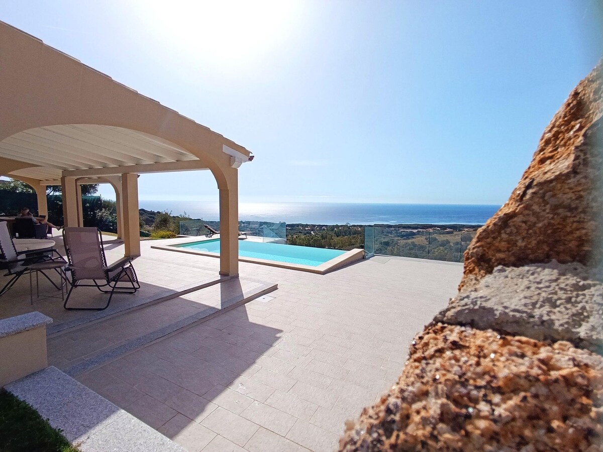 The image depicts a villa's spacious terrace featuring a pool with a view of the sea and landscape. A shaded seating area consists of several lounge chairs, with sunlight illuminating the space under a clear blue sky. A stone wall is visible in the foreground.