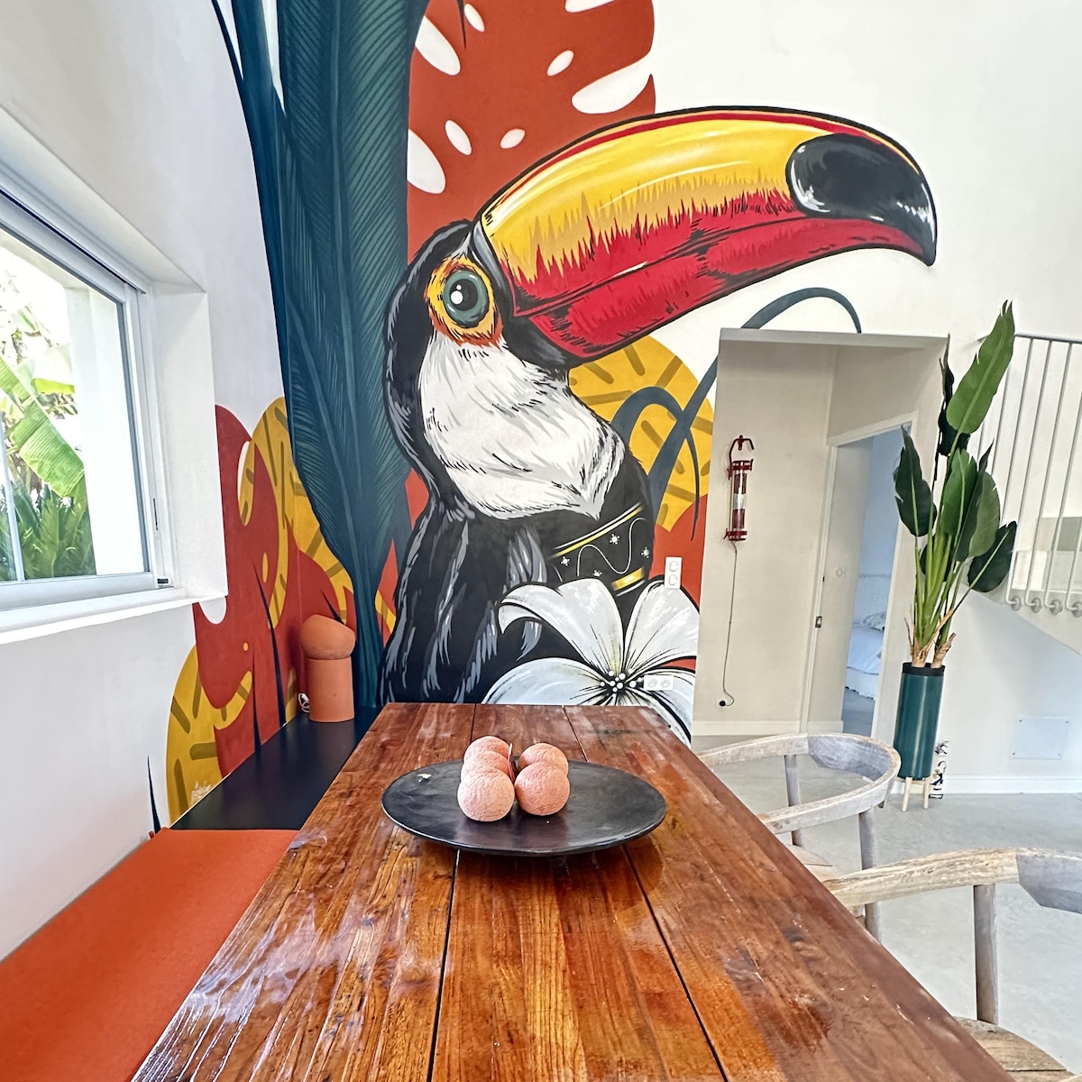 A vibrant wall mural featuring a toucan is displayed next to a wooden dining table. The table is adorned with a round black platter, holding three small items. Natural light enters through a nearby window, illuminating the contemporary space.