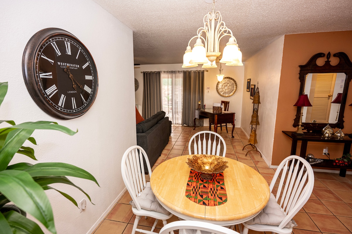 A dining area is presented with a circular table surrounded by white chairs, inviting casual meals. A large clock is mounted on the wall. Soft lighting from a three-bulb chandelier enhances the space, which opens to a living area with a gray sofa.