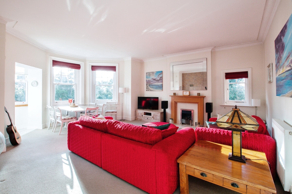 A spacious living area features high ceilings and large windows that provide natural light. Comfortable red sofas are arranged around a wooden coffee table. A decorative lamp sits on the table, and a dining area is visible in the background, with a light-colored table and chairs.