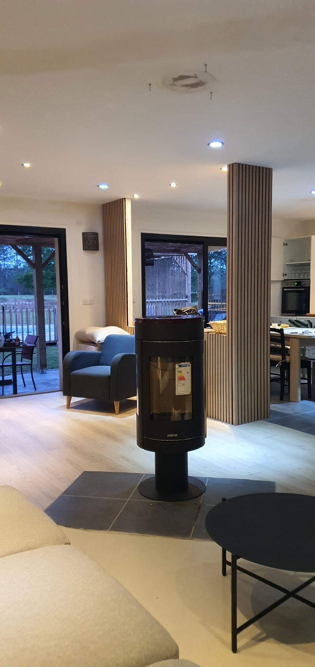 An open living area is highlighted by a modern black stove at the center. Soft seating is arranged around, with large glass doors opening to the outdoor space. Natural light filters through, illuminating the wooden accents and inviting a cozy atmosphere.