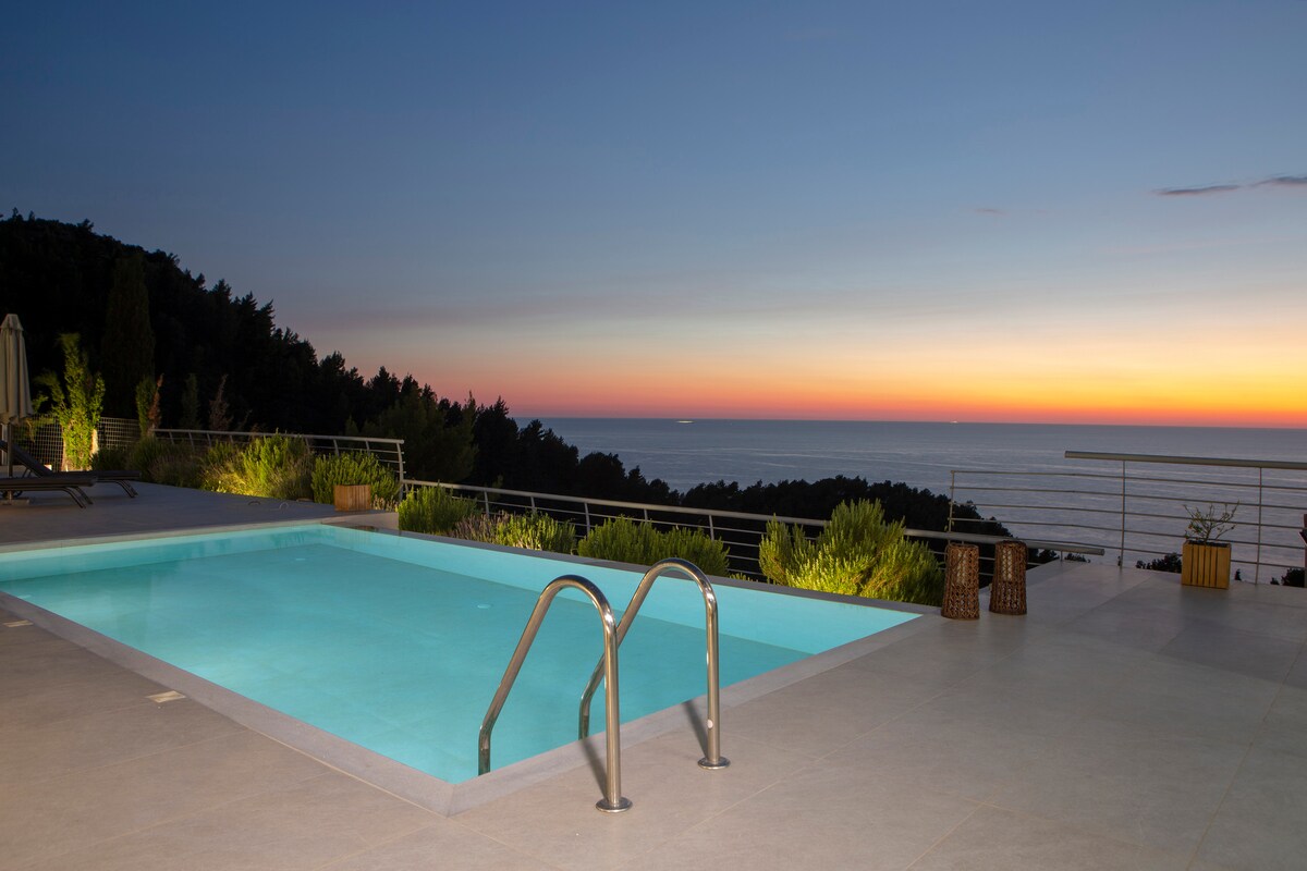 The outdoor area features an infinity pool with clear, calm waters, reflecting the colors of the sunset over the Ionian Sea. A spacious terrace displays greenery surrounding the pool, enhancing the serene atmosphere. The fading light creates a peaceful ambiance in the evening sky.