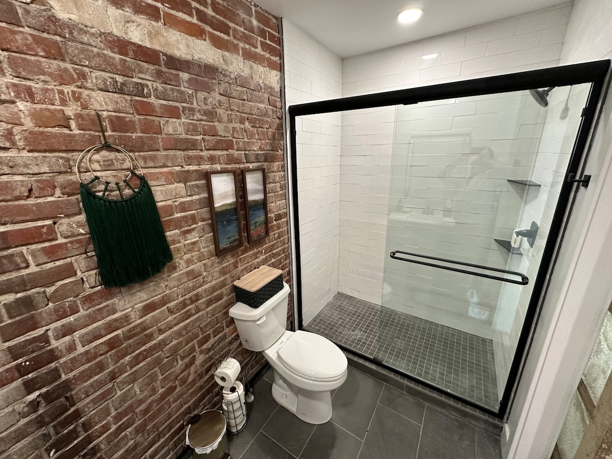 A modern bathroom features a glass-enclosed shower with a tiled floor. Exposed brick walls contrast with sleek white tiles. A toilet and a decorative wooden stool are visible, alongside a framed art piece on the wall and a green wall hanging.