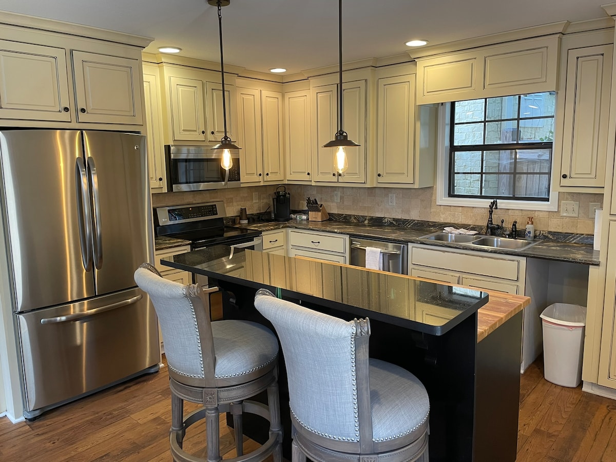 A fully equipped kitchen features warm wood flooring and light cabinets. Stainless steel appliances, including a refrigerator and a stove, are present. An island with seating is highlighted, alongside ample countertop space. Warm lighting from overhead fixtures enhances the functional layout.