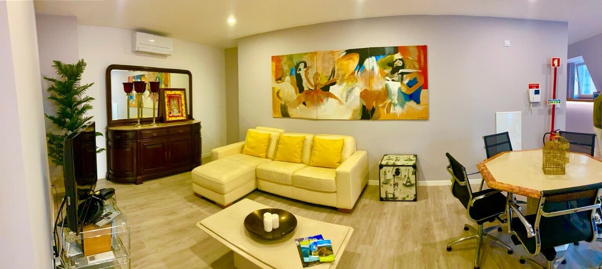 A spacious living area is featured, showcasing a light-colored sectional sofa with yellow cushions. A large abstract painting adorns the wall, while a wooden cabinet and a round dining table with black chairs provide additional furnishings. A small decorative item and magazines are placed on the coffee table.