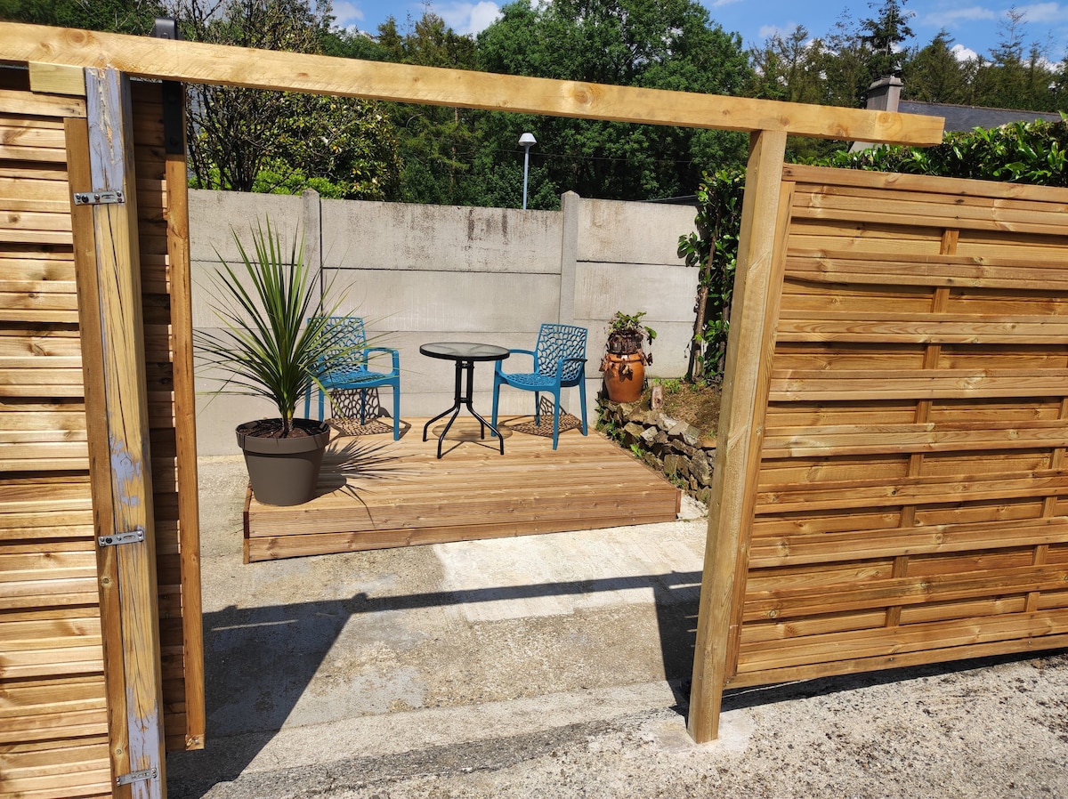 A private outdoor area is accessed through a wooden gate, featuring a small table surrounded by two blue chairs. An ornamental plant is placed on the ground next to the table, and the wooden deck is bordered by stone walls on one side.