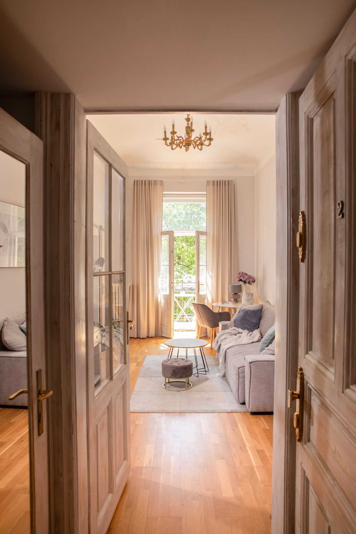 A welcoming entrance leads into a well-lit living area featuring large windows with soft curtains. A comfortable sofa is arranged around a coffee table, inviting relaxation. Light wooden floors enhance the airy ambiance, while a chandelier adds a touch of elegance to the space.