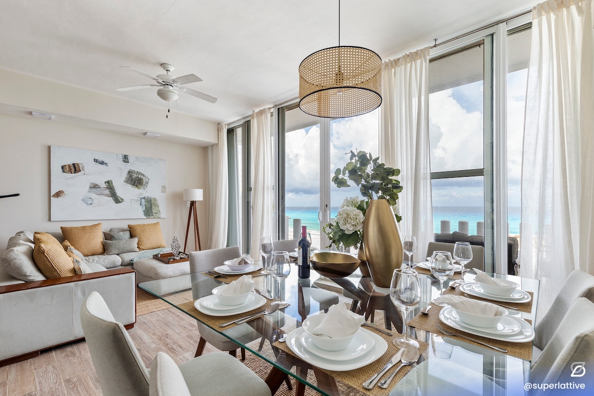 A welcoming living and dining area showcases large windows with views of the sea. A dining table is set for eight, complemented by stylish dishware. A cozy sofa is positioned nearby, adorned with neutral-toned cushions. Natural light fills the space, enhancing the tranquil atmosphere.