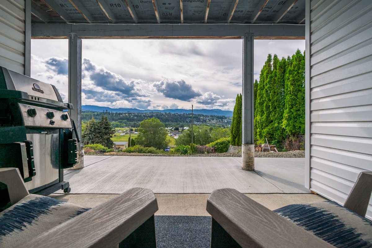 A covered patio area features two cozy chairs positioned for viewing the expansive valley landscape. A BBQ grill stands nearby, and a scenic backdrop of rolling hills and cloudy skies extends beyond the patio, inviting guests to relax and enjoy the outdoor environment.
