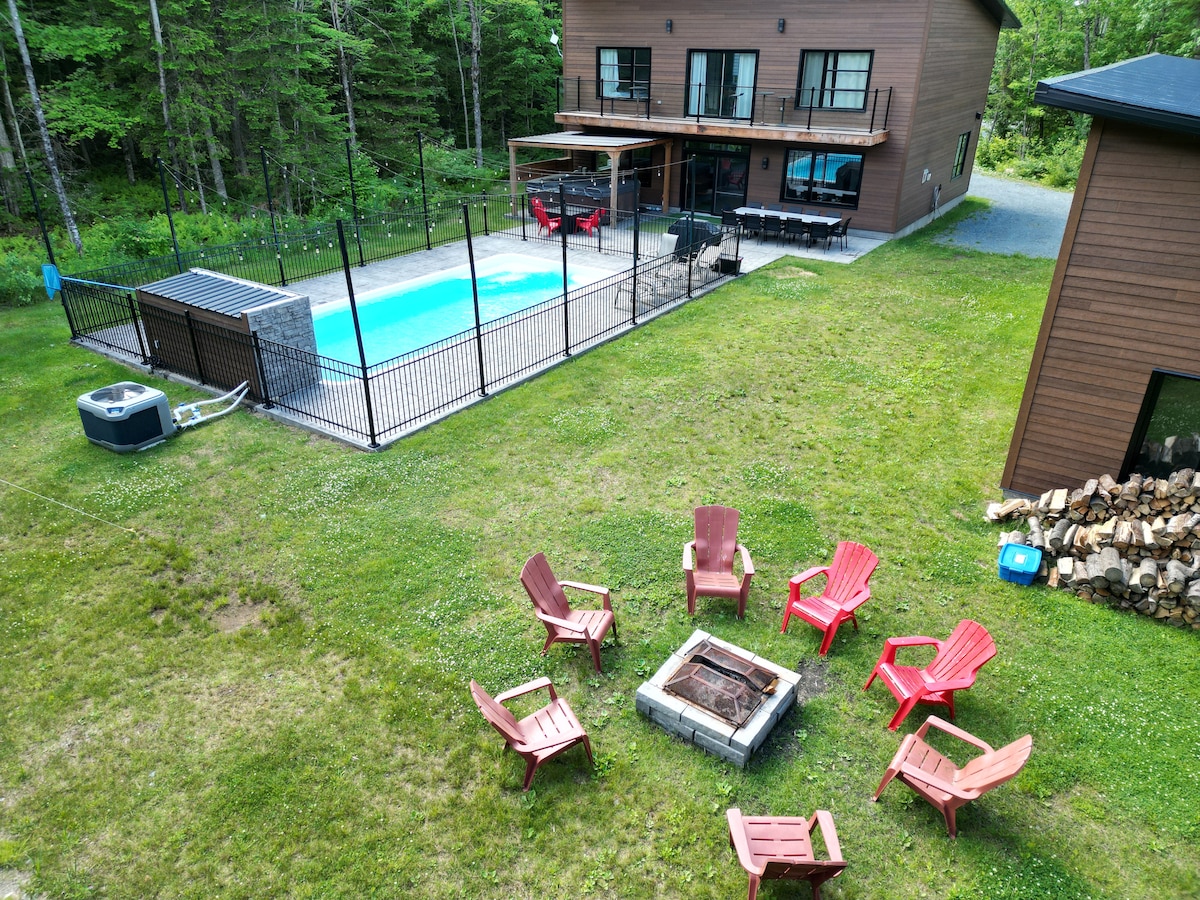 An outdoor area features a neatly arranged circle of red Adirondack chairs surrounding a fire pit, situated on a grassy lawn. A fenced-in swimming pool is visible, alongside a spacious deck equipped with seating, while a wooded backdrop provides a natural setting.