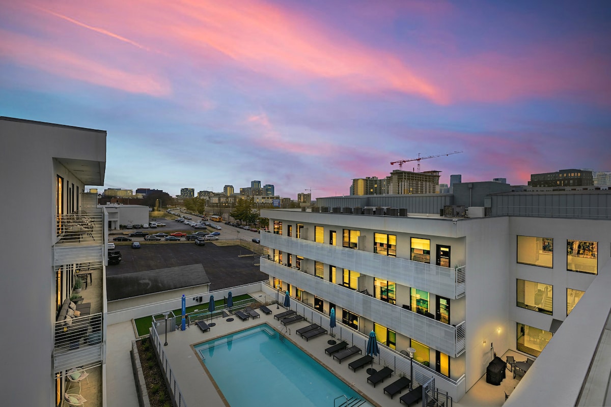 The image captures a modern condo complex at dusk, showcasing a swimming pool surrounded by lounge chairs. A colorful sunset casts hues of pink and purple across the sky, while city skyline elements appear in the background. The structure highlights a contemporary design with balconies.