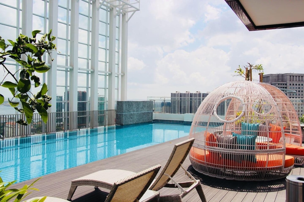 A rooftop pool area features a striking design with a circular lounge surrounded by vibrant cushions. Sun loungers are placed nearby, and tall glass windows reflect sunlight, creating a bright and open environment. A serene water surface enhances the overall ambiance.
