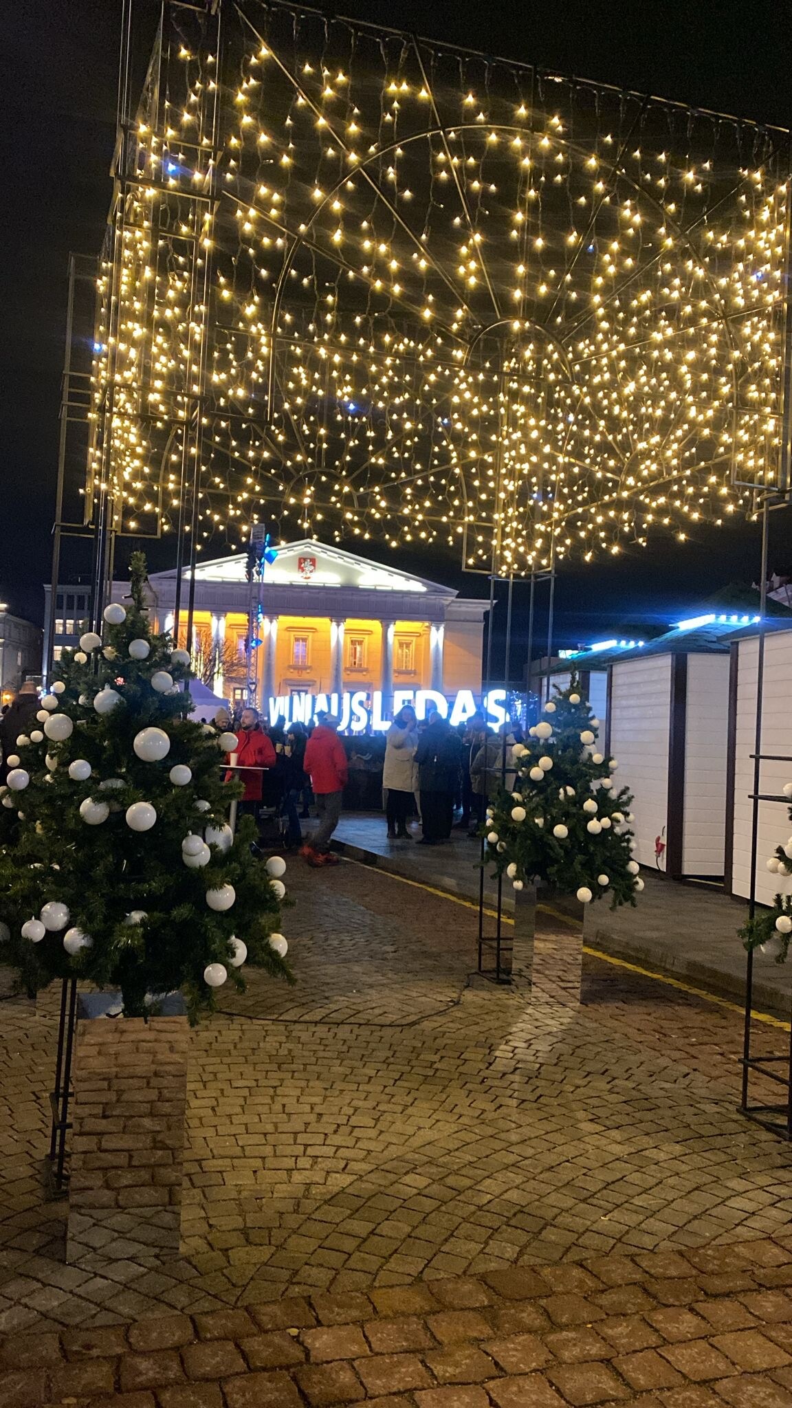 The image displays a festive atmosphere with sparkling lights above a cobblestone pathway. Evergreen trees adorned with white ornaments are positioned along the walkway, leading to a brightly lit building in the background, which features welcoming signage.