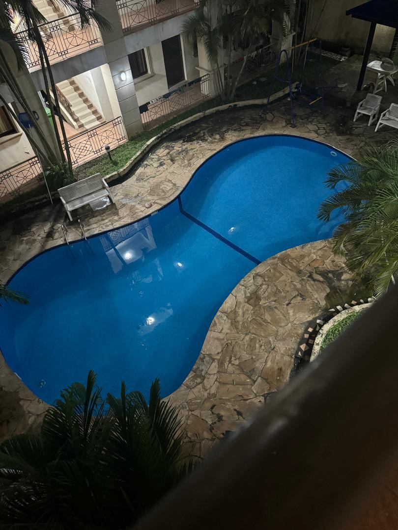 A clear blue swimming pool is framed by natural stone tiles and surrounded by lush greenery. Sun loungers are positioned nearby, offering a relaxing space. The pool's curvaceous design adds to its inviting appearance, with shadows cast from palm fronds creating a serene ambiance.