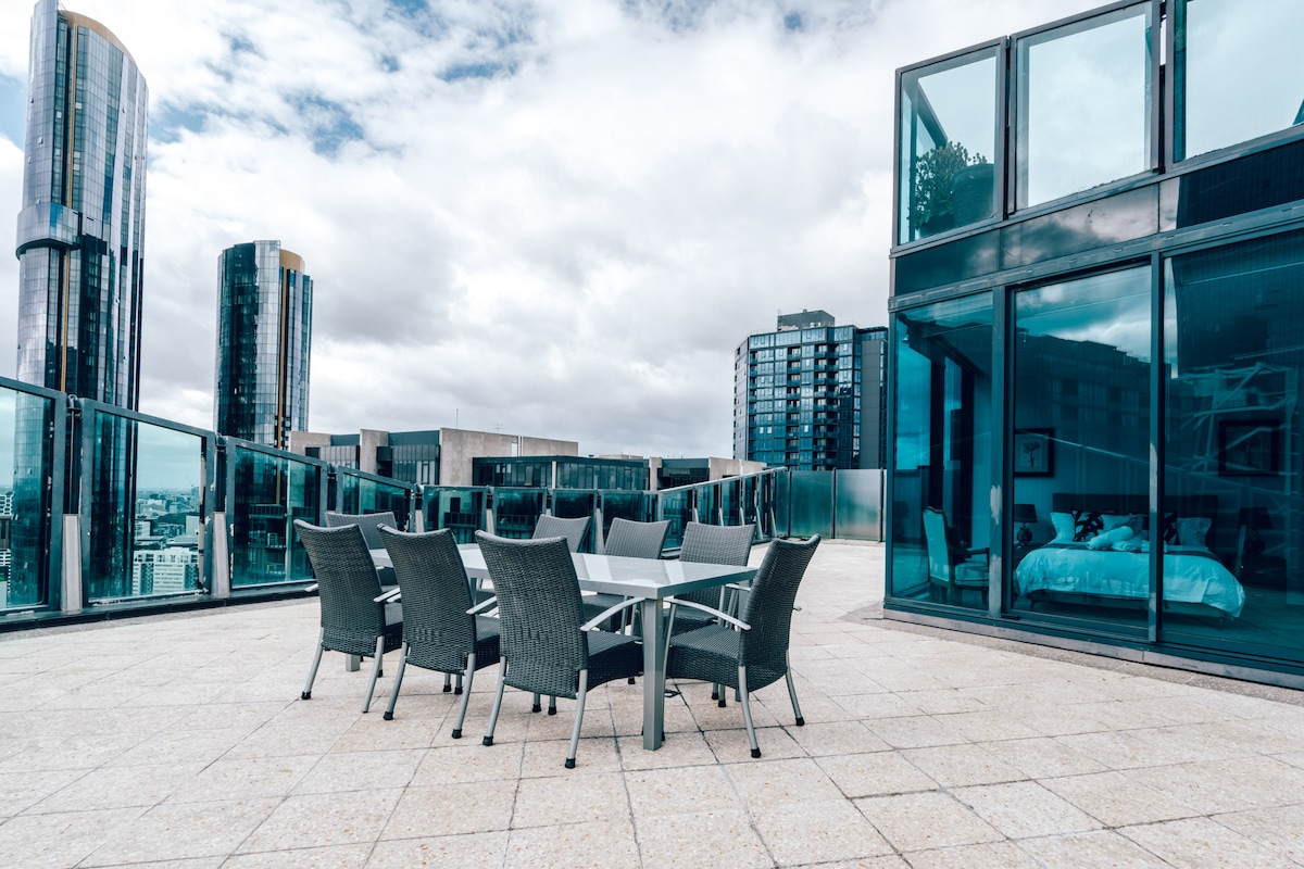 An expansive balcony is featured, furnished with a large dining table surrounded by eight comfortable chairs. The surrounding glass railing offers unobstructed views of the city skyline. A portion of the penthouse interior is visible through floor-to-ceiling glass doors, revealing a well-appointed sleeping area.