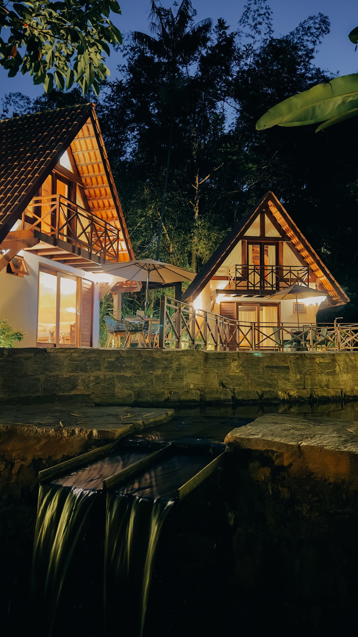 Two charming chalets are visible under the evening sky, featuring steeply pitched roofs and large windows. A stone pathway leads to a sitting area with an umbrella. Soft lighting from within the chalets creates a warm glow, enhancing the natural surroundings.