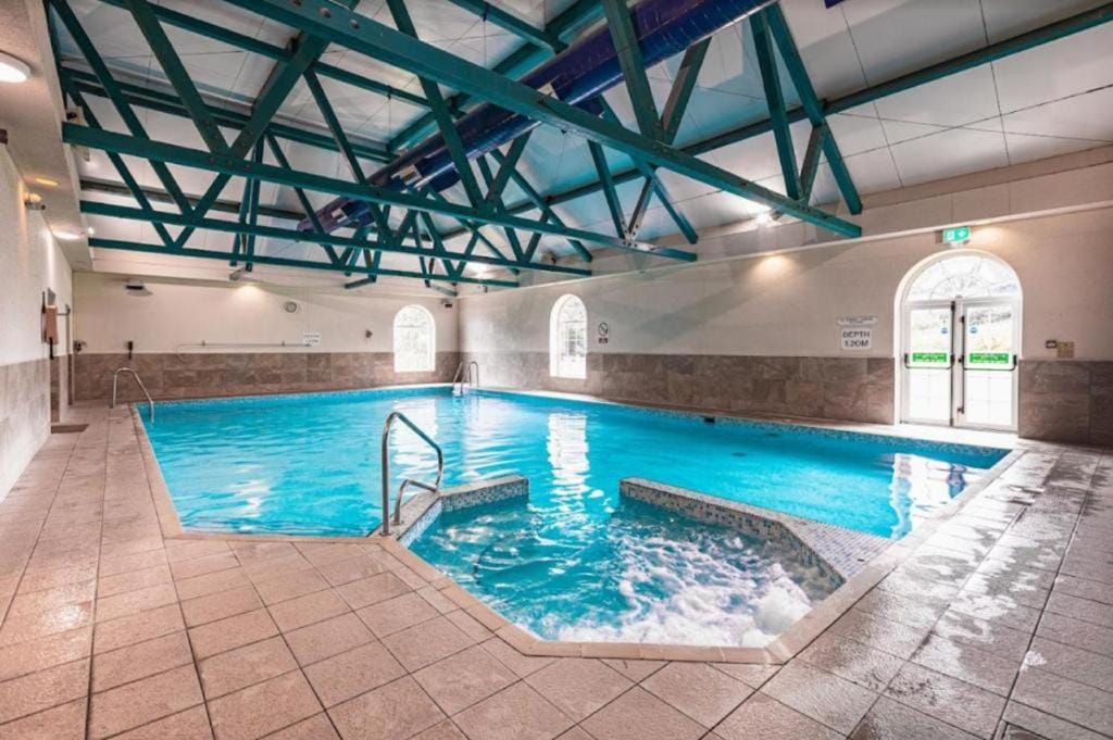 An indoor swimming pool is seen, featuring modern turquoise beams and a bright blue water surface. The area includes a hot tub and large windows allowing natural light, with tiled flooring surrounding the pool, creating an inviting ambiance for relaxation.