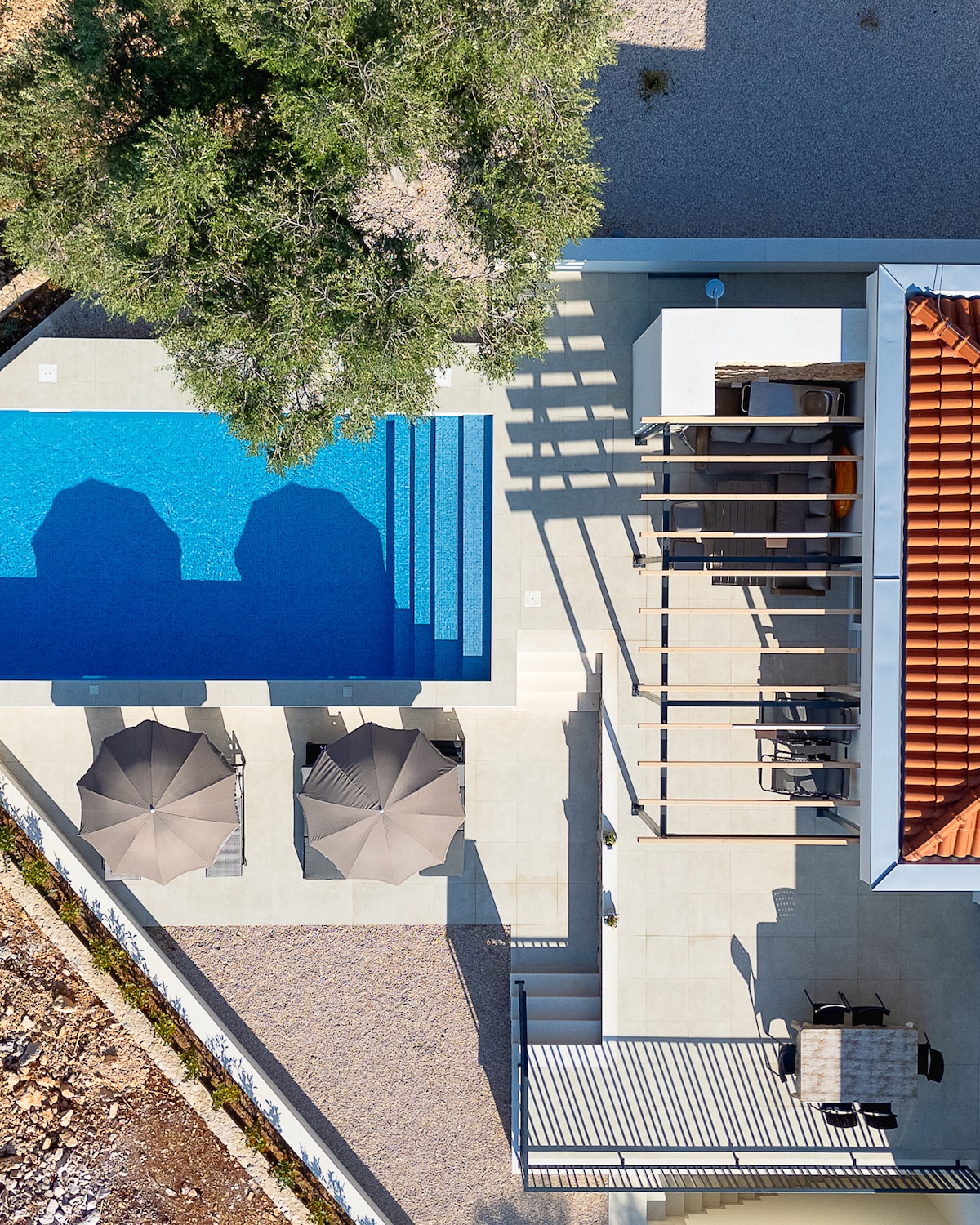 An aerial view captures a private pool surrounded by sun loungers under umbrellas. A shaded terrace is visible adjacent to the pool, along with a dining area. The villa's modern architecture is complemented by natural landscaping, emphasizing outdoor leisure space.