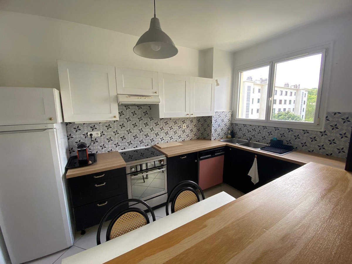 A modern kitchen is featured, showcasing a combination of white and black cabinetry complemented by wooden countertops. Large windows allow natural light to fill the space. A dining area with four chairs is positioned alongside, creating a functional layout for meal preparation and dining.